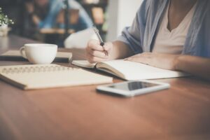 female writing on wood desk in a notebook with a cup and a phone nearby